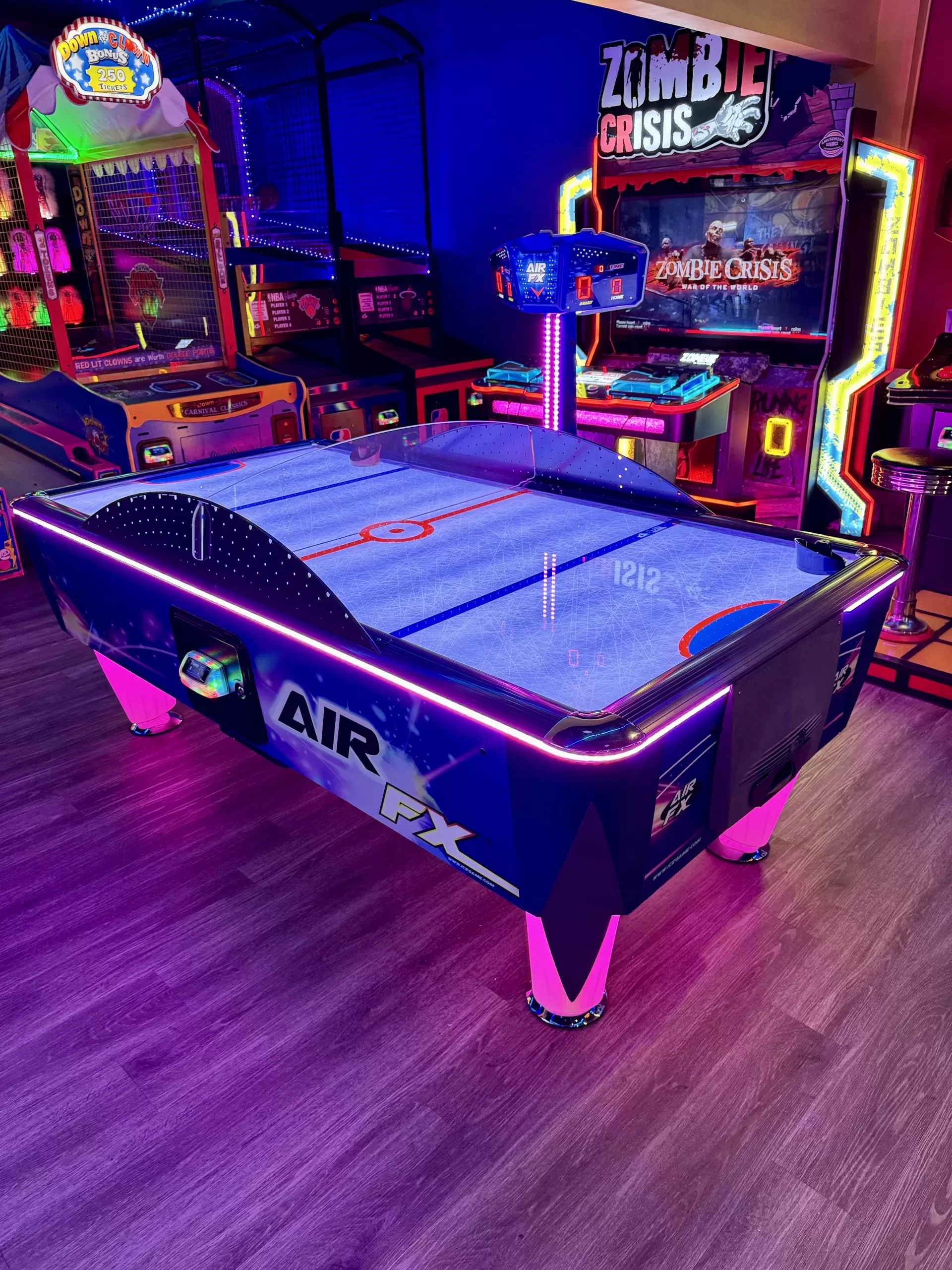 Air hockey table glowing with neon lights in an arcade, surrounded by colorful games including Zombie Crisis.