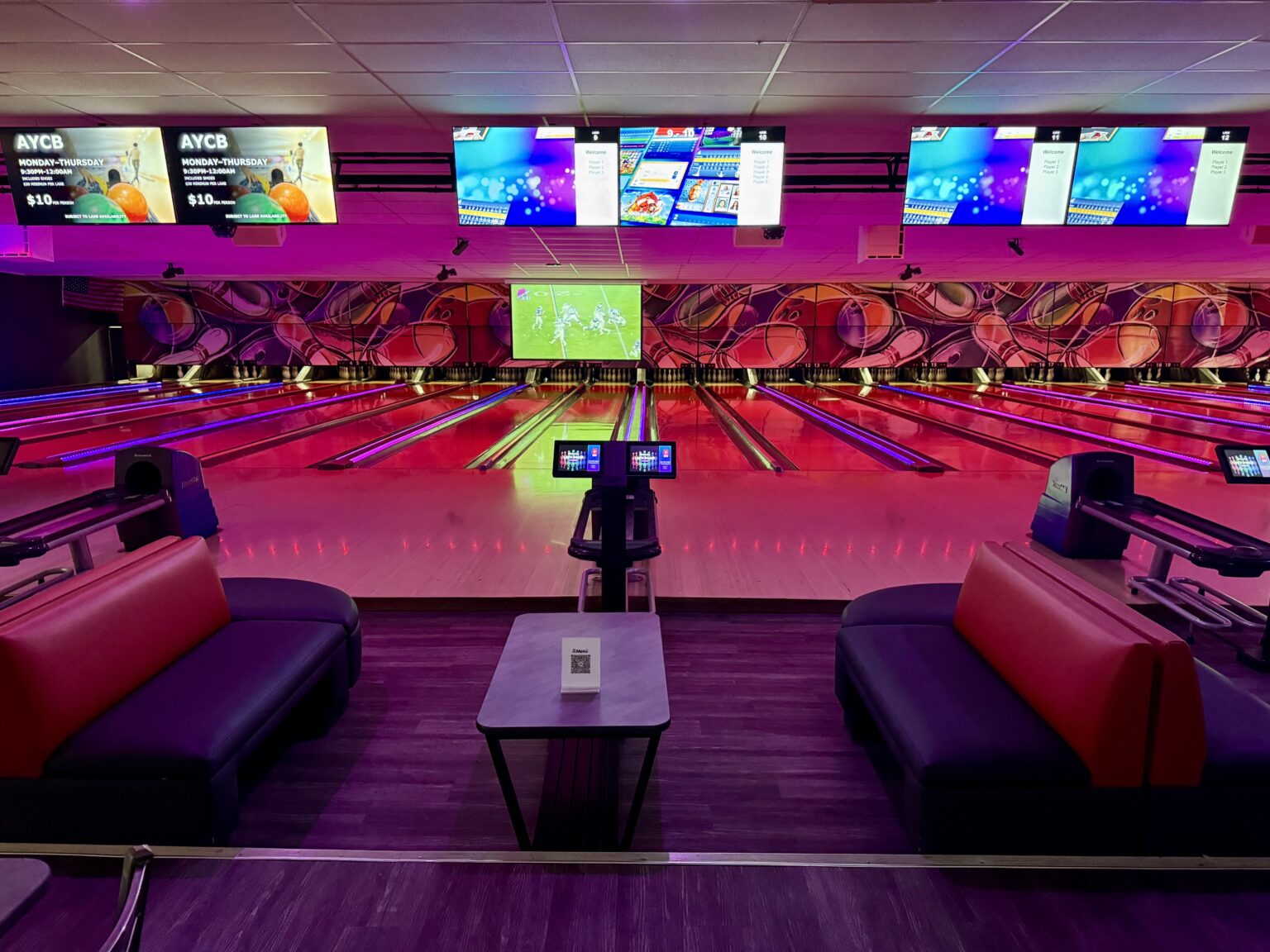 A modern bowling alley with vibrant neon lighting, comfortable lounge seating, and multiple digital screens displaying bowling scores and a sports game. The lanes are illuminated with colorful lights, creating an exciting and immersive atmosphere.