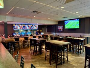 Modern sports bar with high-top tables, a fully stocked bar, and multiple large-screen TVs displaying live games at Strikers Bowling.